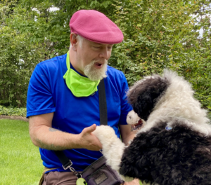 A person with a white goatee, wearing colorful clothes, is holding the paw of a very fluffy curly haired dog, and talking to them.